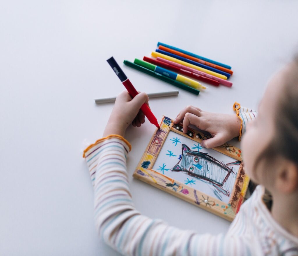 A girl sits at a table and draws, decorates a frame for a photo with drawing, children creativity